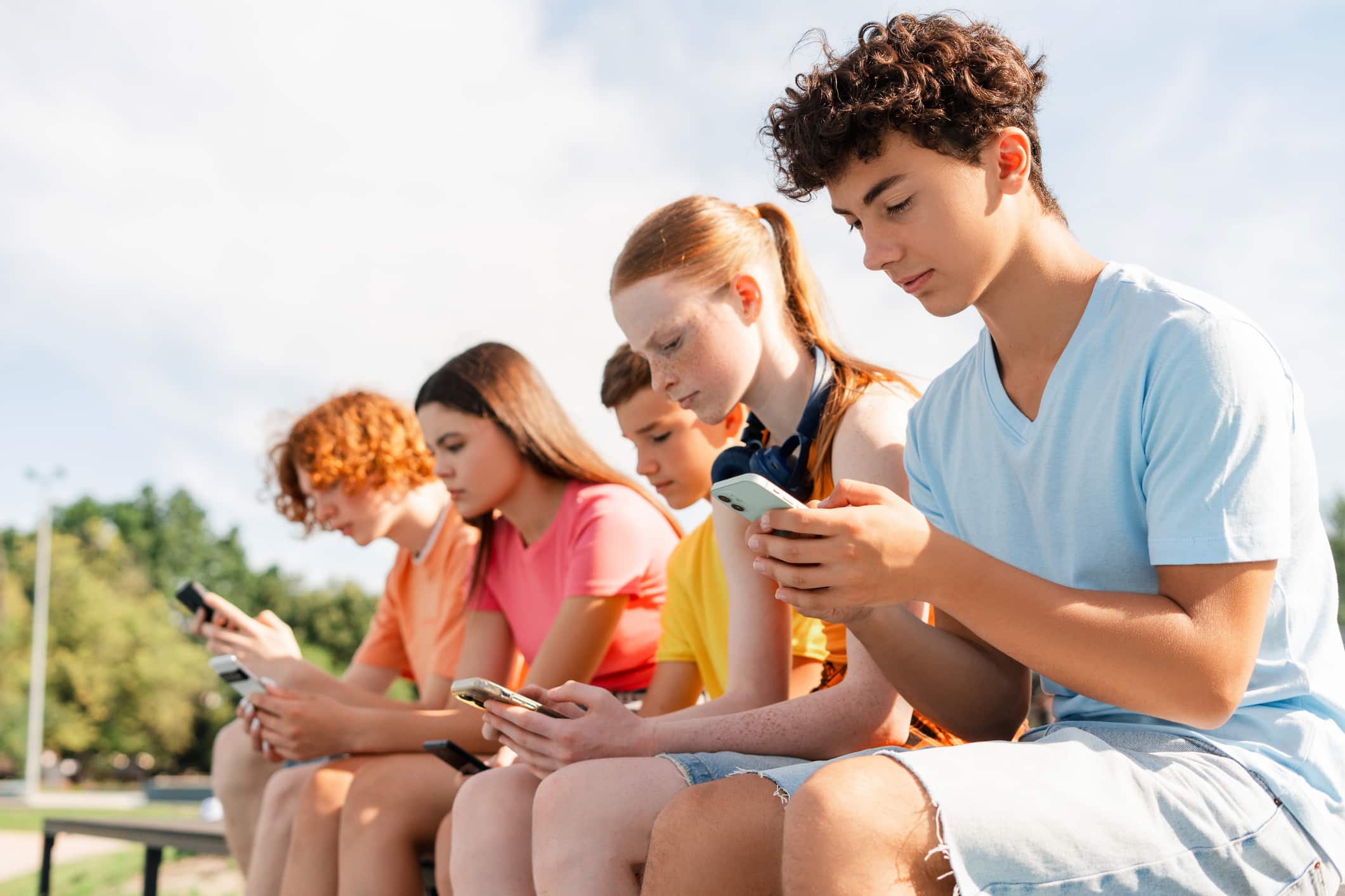 Group of teenagers sitting together outdoors looking at their phones, representing how family vacations with teens can encourage real connection beyond screens.
